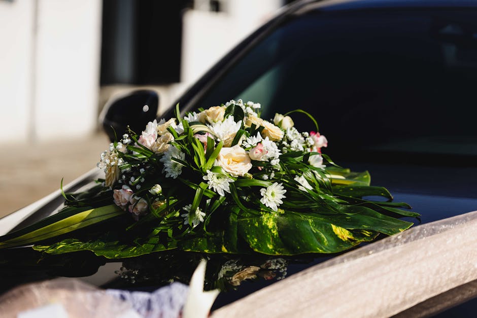 A floral arrangement of fresh white roses, small white daisies, and delicate pink-tinted flowers, all nestled among lush green foliage with glossy leaves, is displayed on the hood of a black car. The 