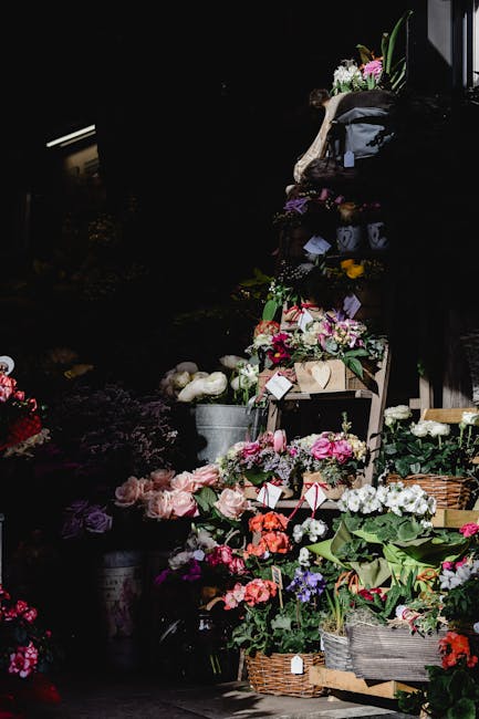 A display of various fresh flowers arranged in baskets and wooden crates, featuring pink roses, white peonies, purple irises, and red anemones, set on a dark outdoor background with ambient lighting h
