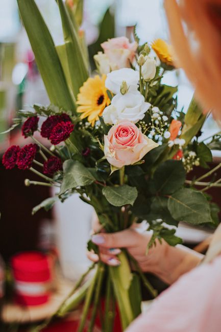 A hand holding a fresh floral bouquet featuring a variety of flowers, including a soft pink rose at the center, surrounded by small white blooms, bright yellow daisies, and deep red carnations. The bo